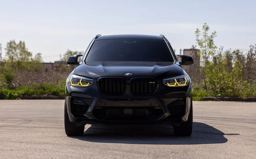 A black BMW X4 M SUV photographed from the front on an outdoor paved surface with trees and shrubs in the background. The car has distinctive yellow LED headlights and a large black kidney grille.