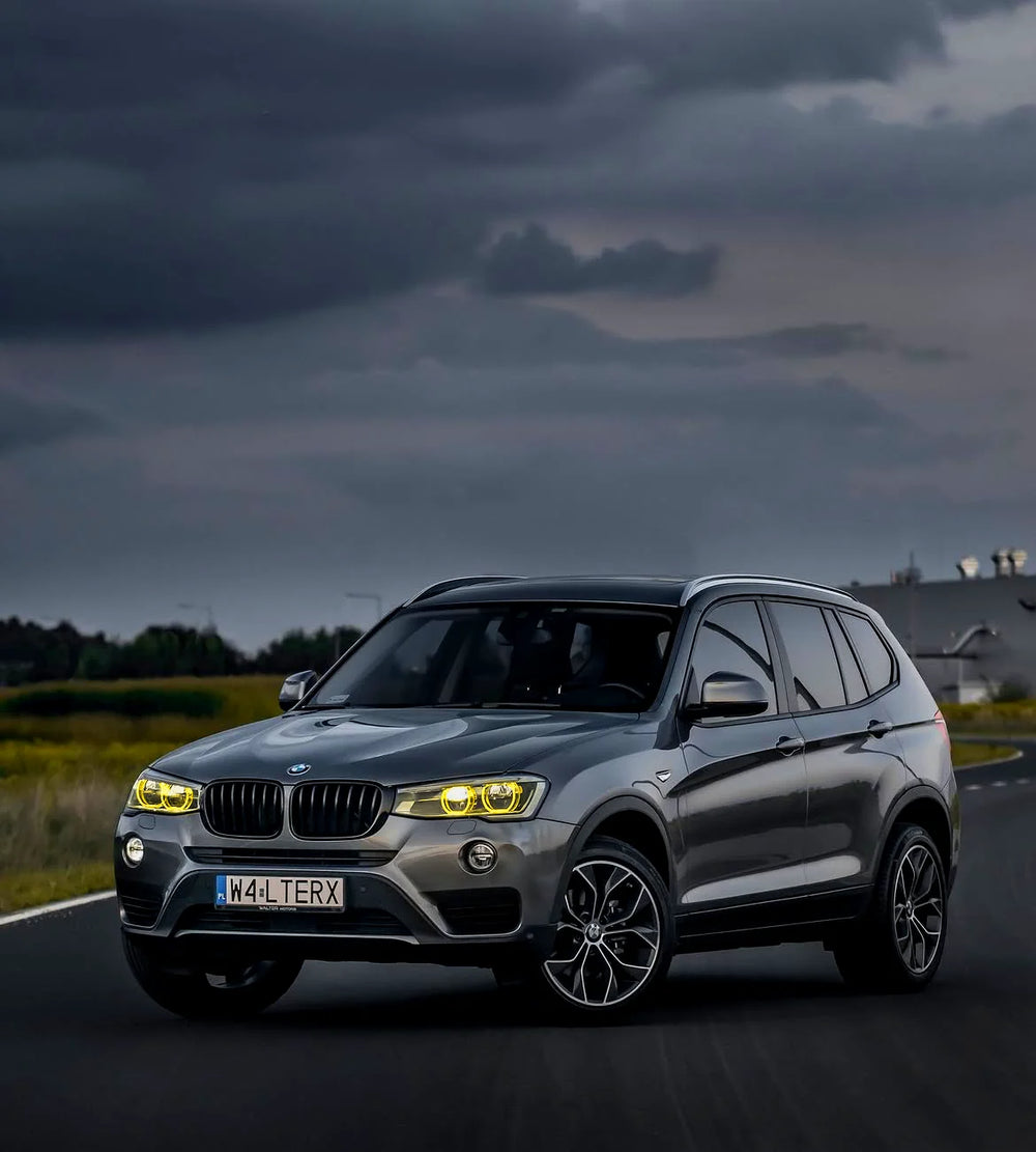 A metallic gray BMW X3 SUV is captured from a dynamic three-quarter angle on a dark road under a dramatic, cloudy sky.