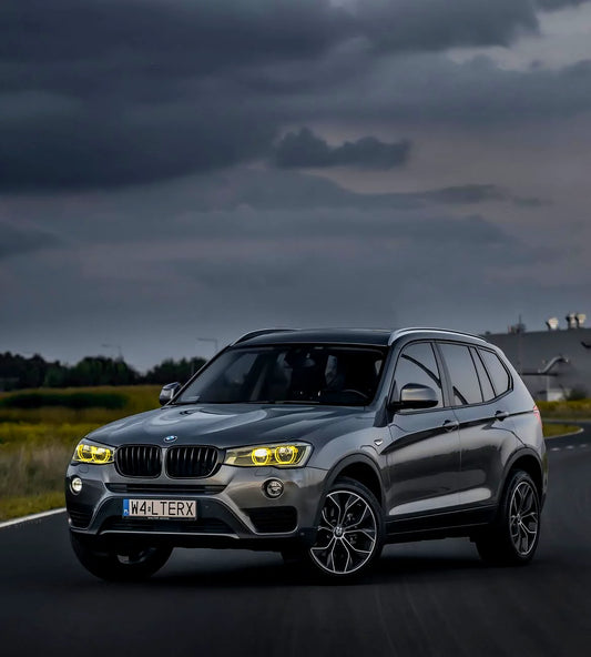 A metallic gray BMW X3 SUV is captured from a dynamic three-quarter angle on a dark road under a dramatic, cloudy sky.